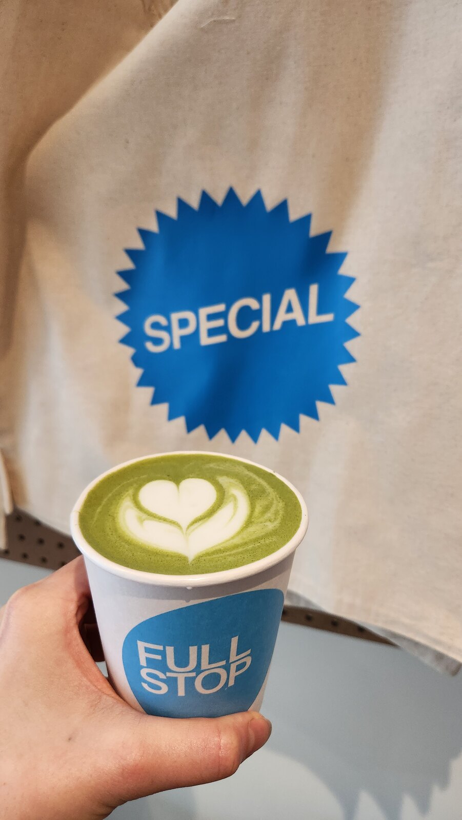 A hand holding a green latte with heart-shaped latte art in a Full Stop cup.