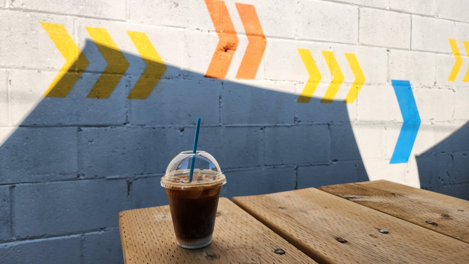 An iced coffee on a wooden patio table beside a colourful painted wall.