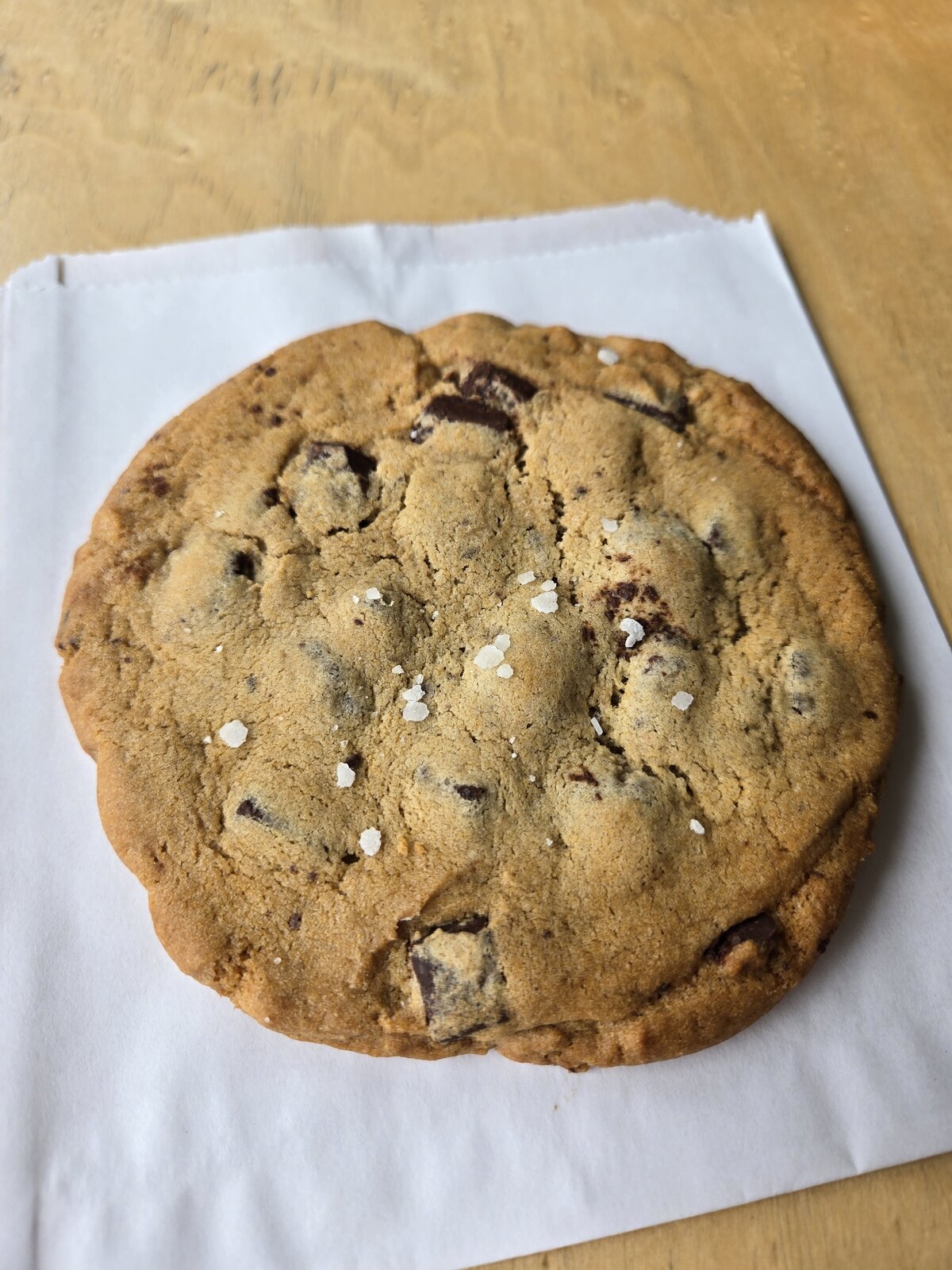 A chocolate chip cookie on white paper on a wooden table.