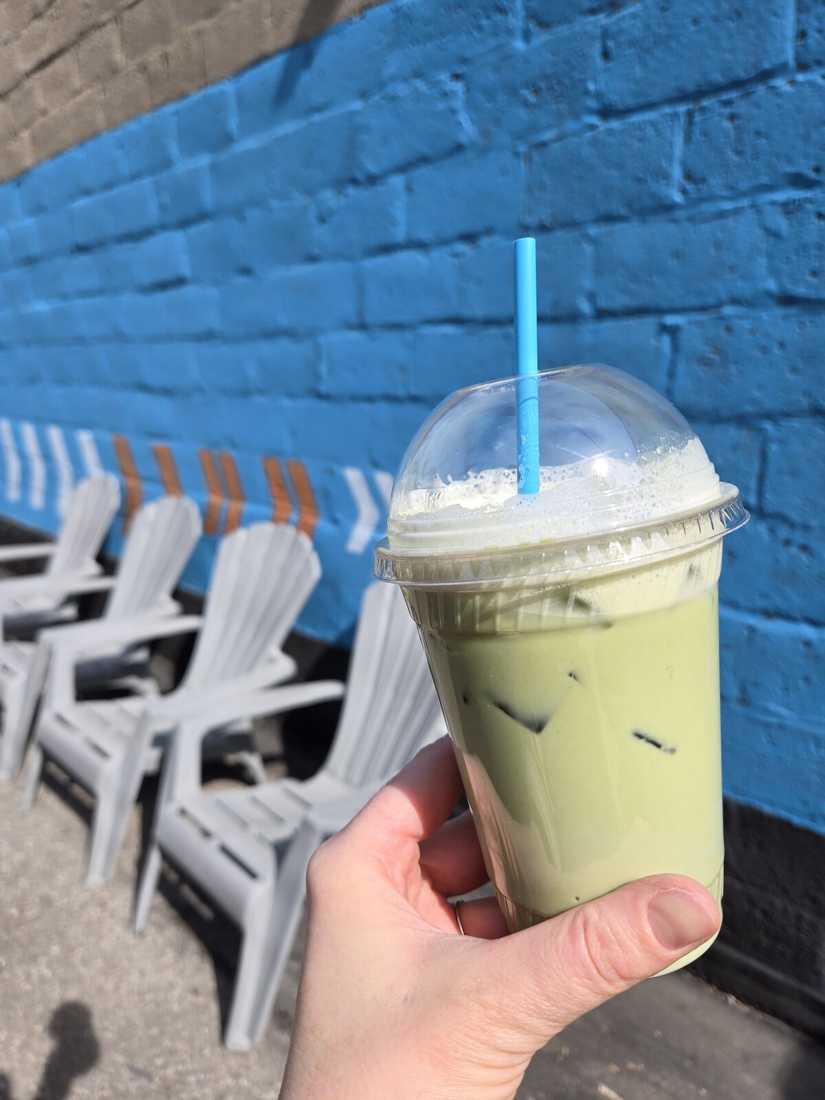 A hand holding an iced green latte beside a blue brick patio wall.