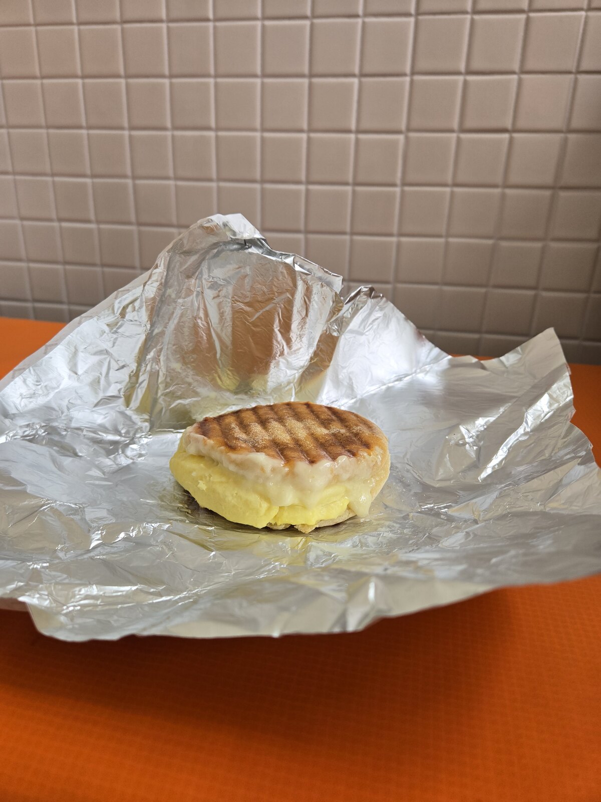 A grilled breakfast sandwich in an open foil wrapper on an orange table.